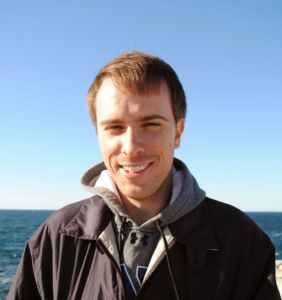 Brian DeConinck, a white man in his mid-30s. He is wearing a sweatshirt and a jacket, and is standing in front of the ocean on a clear day.